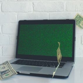 Laptop computer with green computer code running across screen. Dollar bills fall around the laptop.
