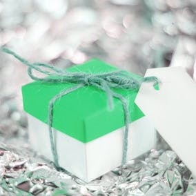 A green and white gift box that has a blank label tied to it with twine. Gift box sits on a silver, reflective surface.  