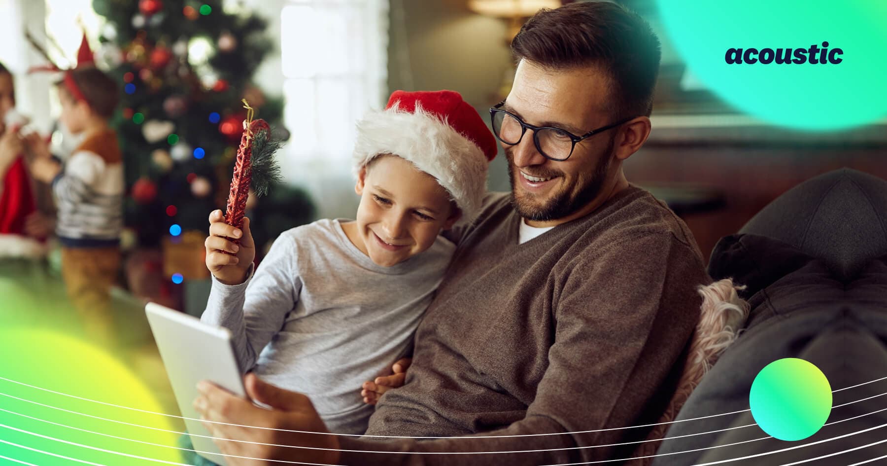 Father and son wearing a Santa hat looking at a tablet together on a couch beside a decorated Christmas tree