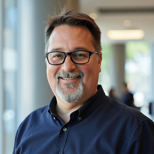 Headshot of a smiling man with glasses and a salt-and-pepper beard wearing a dark button-up shirt, professional services team member