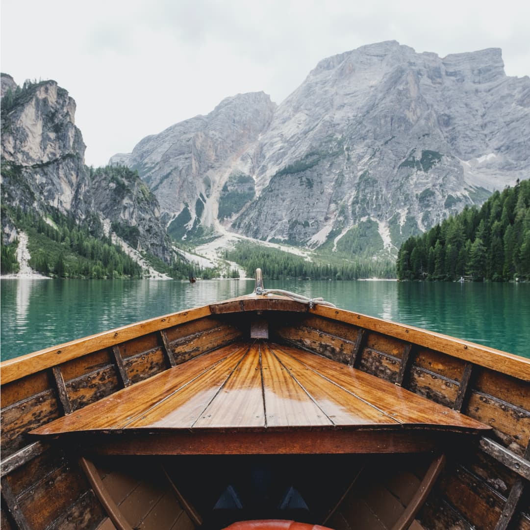 Bow of a wooden boat on a smooth lake surrounded by mountains and trees.