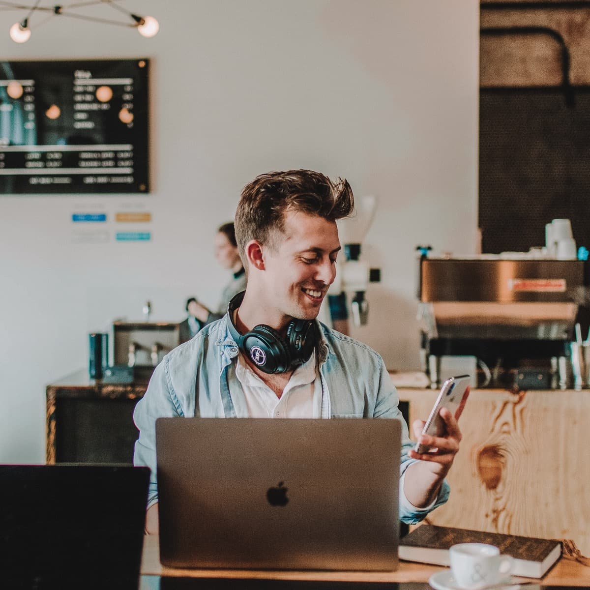 Image of a person sitting in a coffee shop with a laptop, coffee, and book in front of him. He's smiling and looking at his phone.